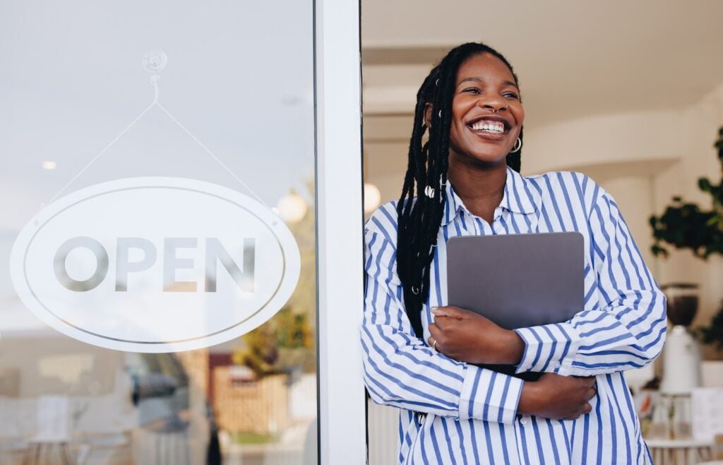 Successful young cafe owner standing next to an open sign at the entrance of her new restaurant. Female small business owner smiling happily while holding a digital tablet in her coffee shop.