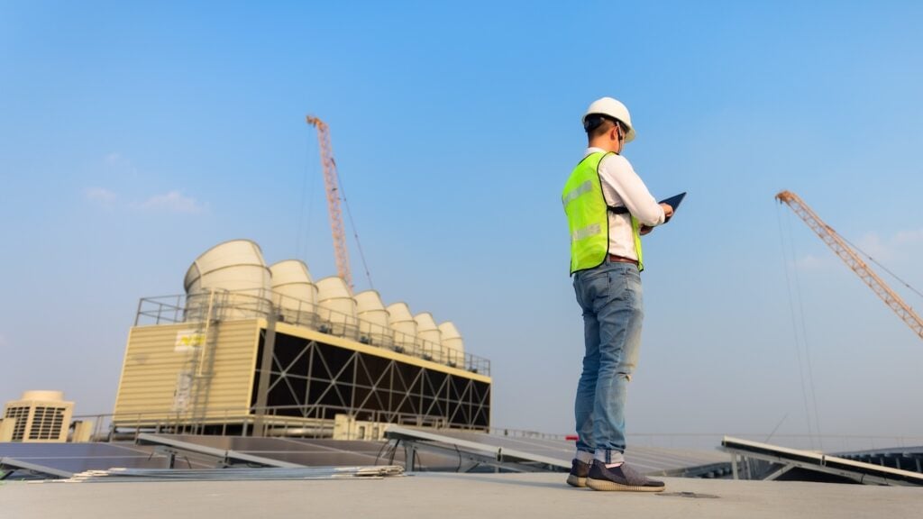 Engineer holding tablet is checking the cooling tower and solar cell on the roof of the building to be in good condition.