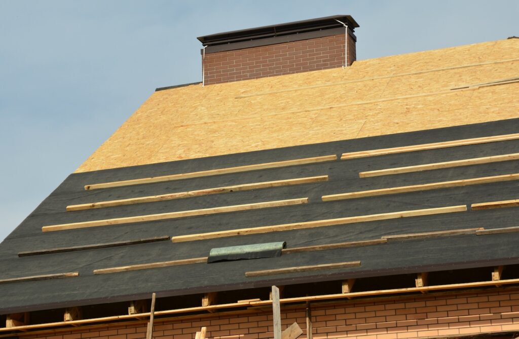 A close-up of a gable incomplete roof with a broad brick chimney, sheathed with plywood and with waterproof and water-resistant underlayment installed before roofing asphalt shingles installation.