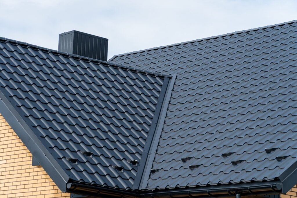 Dark grey metal roof tiles on a modern house with chimney and light brick wall.