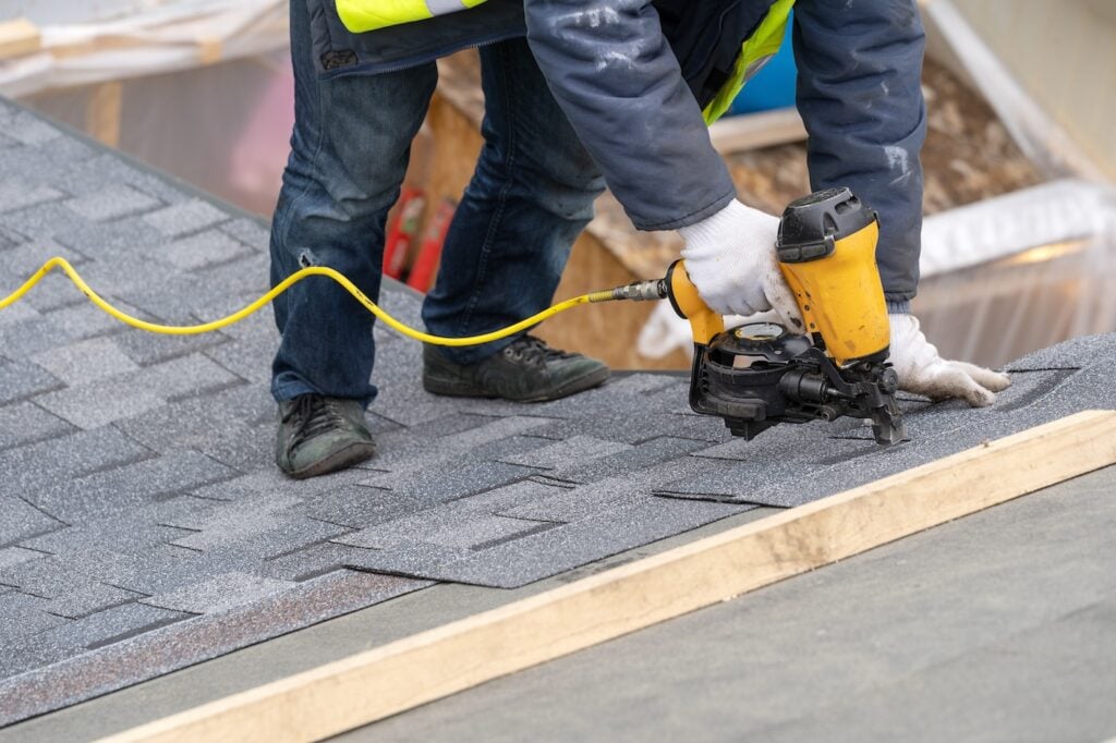 Close up and real photo of professional roofer worker in uniform work wear using air or pneumatic nail gun and installing asphalt or bitumen shingle on top of the new roof under construction residential building