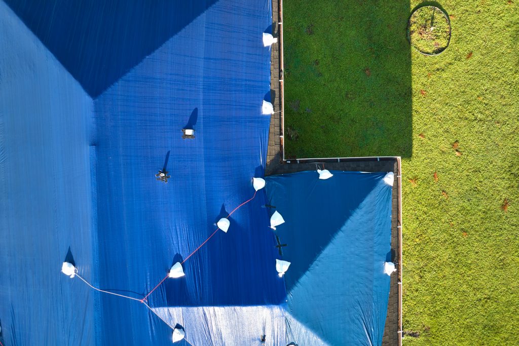Hurricane Ian damaged house rooftop covered with protective plastic tarp against rain water leaking until replacement of asphalt shingles. Aftermath of natural disaster.